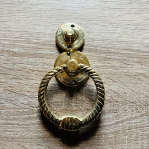 Può includere: Un battente per porta in ottone lucido caratterizzato da una piastra di montaggio circolare con un motivo inciso a forma di occhio e una piastra secondaria fissata a un pesante anello a corda intrecciata che pende da un perno centrale.