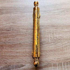 May include: A cylindrical gold-coloured metal door handle featuring an intricate engraved floral pattern along its length. The handle has a smooth, polished finish with rounded, spherical finials at both the top and bottom ends of the piece.