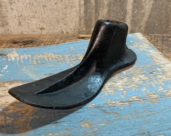 May include: A black metal shoe last, used by shoemakers to shape and form shoes. The last is resting on a piece of weathered wood with blue paint.