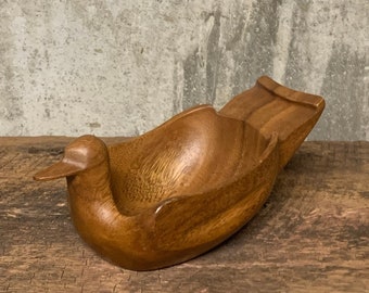 May include: A wooden bowl carved in the shape of a bird. The bowl is brown and has a smooth finish. It is sitting on a wooden surface.