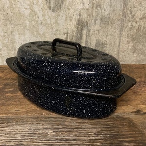 May include: A black speckled enamel roasting pan with a lid and a handle. The oval-shaped pan has a dark speckled pattern and is sitting on a wooden surface. The pan has two side handles.