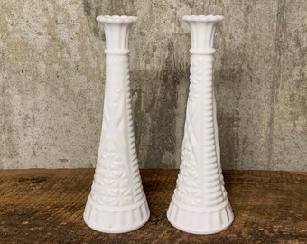 May include: Two white milk glass bud vases with a textured design. The vases are tall and slender with a flared top.
