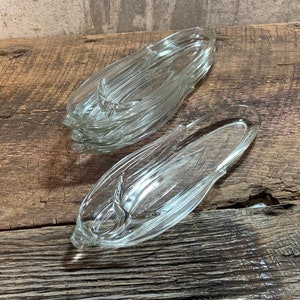 May include: Two clear glass corn on the cob dishes. The dishes are shaped like corn on the cob and have a ribbed texture. The dishes are sitting on a wooden surface.