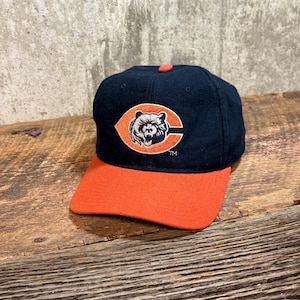 Puede incluir: Una gorra de béisbol azul marino con visera naranja y un logotipo central bordado de una cabeza de oso dentro de una letra C naranja. La gorra tiene un pequeño botón naranja en la parte superior central y las letras TM junto al logo.