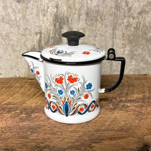 May include: A white enamel teapot featuring a black handle and lid knob, decorated with a colorful floral pattern in red, blue, and green. The design includes stylized flowers and scrolling vines around the body and lid, with a black rim.