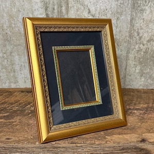 Peut inclure: Un cadre photo doré avec un passe-partout noir et un petit cadre intérieur doré. Le cadre est rectangulaire et présente des détails ornés. Le cadre est posé sur une surface en bois.