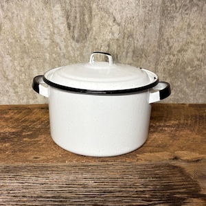 May include: White enamelware pot with a lid and black trim. The pot has two side handles and a handle on the lid. The pot is sitting on a wooden surface. The pot has a speckled design.