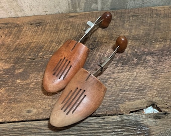 May include: Two wooden shoe trees with metal adjusters and brown wooden handles. The shoe trees are on a rustic wooden surface.