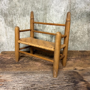 May include: A small, vintage wooden chair with a woven seat. The chair has a light brown finish and a simple design, with four legs and a backrest. The seat is made of woven material. The chair is likely a decorative item.