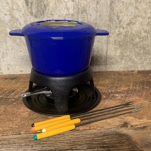 May include: A blue ceramic fondue pot with a black metal stand and four yellow fondue forks. The pot has a round shape and a lid with a hole in the center. The stand has a round base and three legs. The forks have yellow handles and metal prongs.