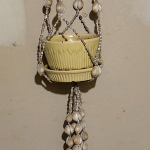 May include: A hanging planter with a yellow ceramic pot and saucer. The planter is suspended by a macrame-style rope adorned with seashells and a cluster of seashells at the bottom. The pot has a ribbed design.