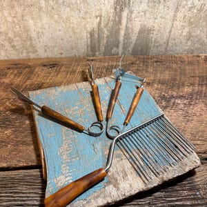 May include: A collection of vintage wool carding tools, including a comb and several hand carders, with brown handles and metal tines. The tools rest on a weathered, blue-painted wooden board, set against a rustic background.