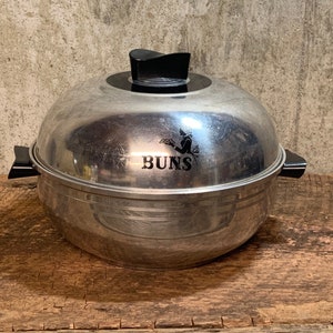 May include: A vintage, stainless steel bun warmer with a black handle and lid knob. The word "BUNS" is printed on the front. The bun warmer is sitting on a wooden surface.