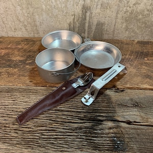 May include: A vintage camping set with three aluminum pots, a folding spoon and fork, and a leather sheath. The set is on a rustic wooden surface.