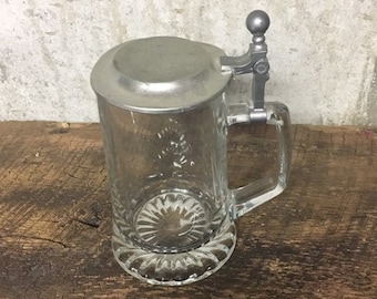 May include: A clear glass beer stein with a pewter lid and handle. The lid has a small knob on top.