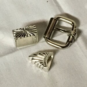 May include: Three silver metal belt buckle pieces. Two are decorative with a sunburst design and the third is a rectangular buckle.