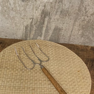 May include: A vintage metal barbecue fork with three prongs and a wooden handle. The fork is on a woven, circular placemat. The background features a textured, grey wall and a wooden surface.
