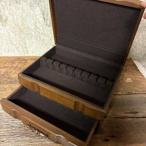 May include: A wooden storage chest with a hinged lid and a pull-out drawer. The interior is lined with dark brown fabric. The top compartment features a slotted insert for organizing items, while the bottom drawer provides an open space.
