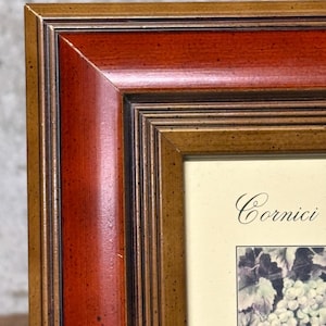 May include: Close-up of a wooden picture frame with a multi-toned finish. The frame features a rich, reddish-brown centre section, bordered by a lighter, golden-brown outer edge. The frame surrounds a picture with the word "Cornici" and a grape illustration.