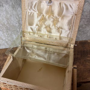 May include: Open wicker sewing box with a clear plastic tray and a cream-colored satin interior. The box has a hinged lid and a woven handle. The interior features a padded satin lining with decorative stitching. The exterior is a light tan color.