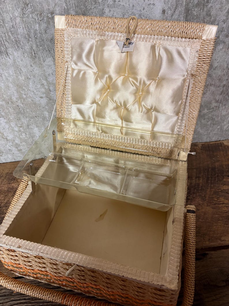May include: Open wicker sewing box with a clear plastic tray and a cream-colored satin interior. The box has a hinged lid and a woven handle. The interior features a padded satin lining with decorative stitching. The exterior is a light tan color.