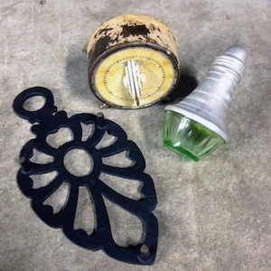 May include: Three vintage items: a black metal trivet with a flower design, a white and yellow kitchen timer with black numbers, and a green glass bottle stopper with a silver top.