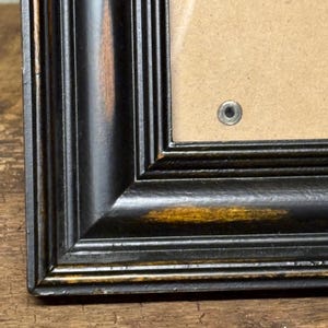 Puede incluir: Un marco de fotos rectangular con acabado negro y desgaste de madera marrón visible en los bordes. El marco presenta un perfil de moldura escalonado de varios niveles e incluye un cierre metálico visible en el fondo de color canela.