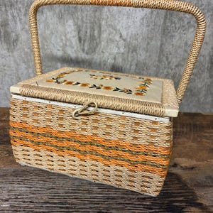 May include: A vintage sewing basket with a woven design in shades of beige and orange. The lid features embroidered floral details. It has a curved handle and a clasp closure. The basket is likely made of wicker or a similar material.