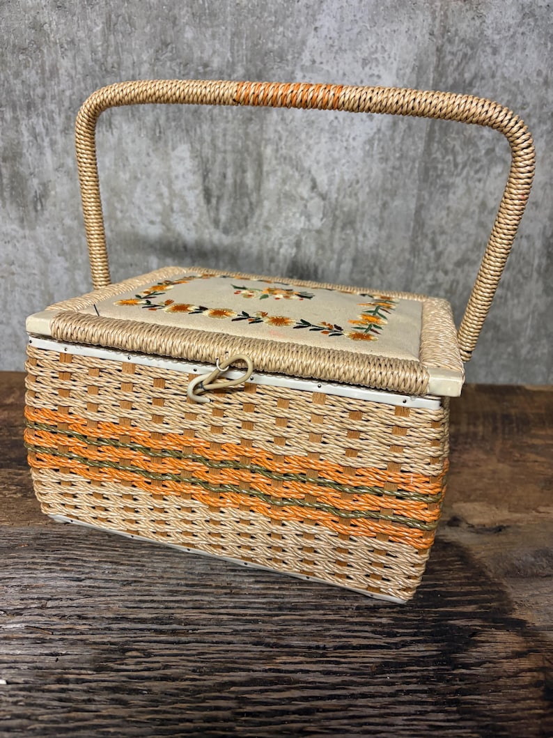 May include: A vintage sewing basket with a woven design in shades of beige and orange. The lid features embroidered floral details. It has a curved handle and a clasp closure. The basket is likely made of wicker or a similar material.