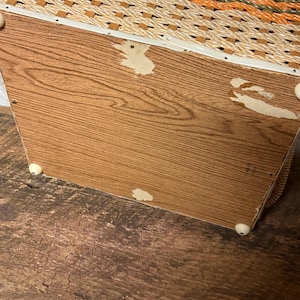 May include: A vintage woven basket with a wooden base. The basket features a woven pattern in shades of beige, orange, and green. The wooden base has a wood grain pattern and is supported by small white feet.