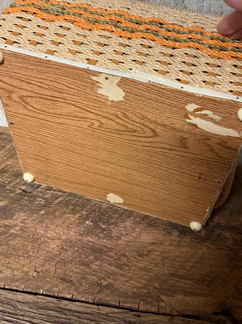 May include: A vintage woven basket with a wooden base. The basket features a woven pattern in shades of beige, orange, and green. The wooden base has a wood grain pattern and is supported by small white feet.