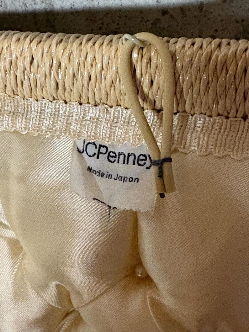 May include: Close-up of a woven straw handbag. The top edge is a tightly woven straw, with a cream-colored satin interior. A tag reads "JCPenney Made in Japan". A beige handle is attached to the top.