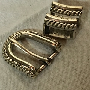 May include: Silver belt buckle with a rope design. The buckle is made of metal and has a polished finish.