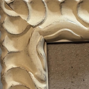 Könnte beinhalten: Nahaufnahme eines cremefarbenen Bilderrahmens mit einem Used-Look. Der Rahmen hat ein wellenförmiges, strukturiertes Design entlang der Kanten, mit dunklen Akzenten in den Rillen. Der Rahmen ist aus Holz und hat eine neutrale, Vintage-Ästhetik.