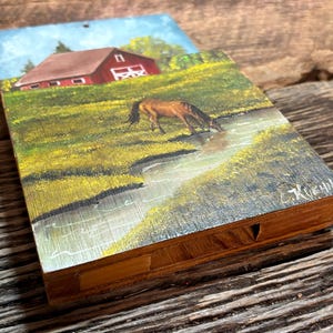 May include: A wooden art piece featuring a painting of a brown horse drinking from a stream in a grassy field. A red barn is in the background under a blue sky. The artwork is set on a rustic wooden surface.