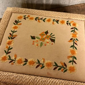 May include: A rectangular, woven wicker box with a beige fabric top. The fabric is embroidered with a floral design featuring yellow flowers, green leaves, and orange accents. The box has a woven border.