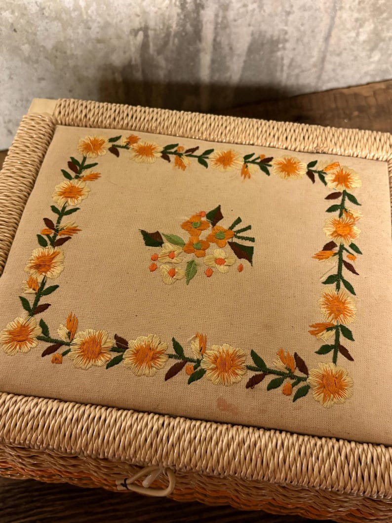 May include: A rectangular, woven wicker box with a beige fabric top. The fabric is embroidered with a floral design featuring yellow flowers, green leaves, and orange accents. The box has a woven border.