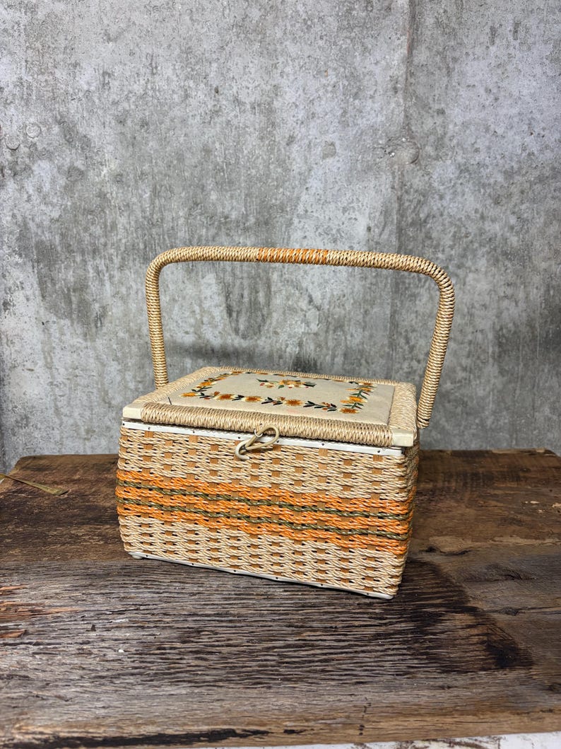 May include: A vintage sewing basket with a woven wicker design in beige and orange. The rectangular box has a hinged lid with a floral embroidered design and a metal clasp. A woven handle arches over the top.
