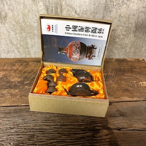 Puede incluir: Un juego en caja de China Cinzhou Clay Pottery Arts. La caja es beige con un interior dorado y contiene una pequeña tetera, varias tazas y un jarrón decorativo. La tapa de la caja presenta una imagen de un jarrón.
