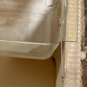 May include: A close-up of a clear plastic container with a beige woven basket. The container has a rectangular shape with a slightly curved edge. The basket has a woven texture and a white string.