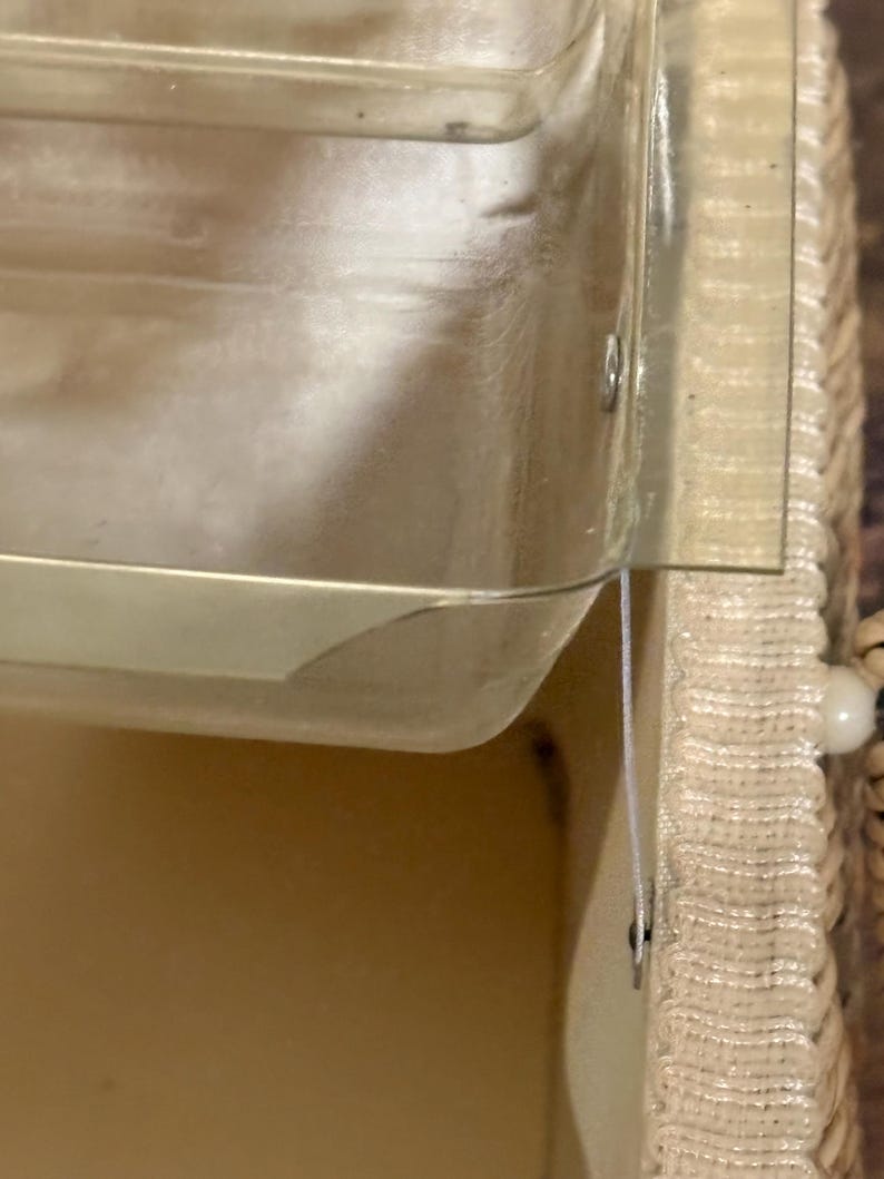 May include: A close-up of a clear plastic container with a beige woven basket. The container has a rectangular shape with a slightly curved edge. The basket has a woven texture and a white string.
