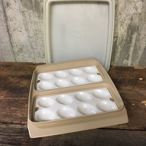 May include: A beige plastic egg container with a matching lid. The container has two rows of oval-shaped indentations to hold eggs. The lid is rectangular. The container is on a wooden surface.