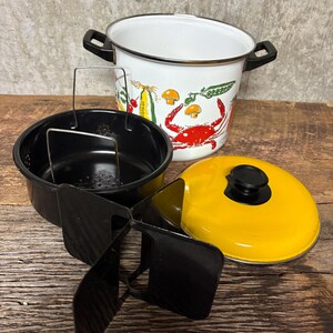 May include: A vintage enamelware cooking pot with a white exterior featuring a colorful crab and vegetable design. Includes a black steamer basket, a yellow lid, and a black metal trivet. The pot has black handles.