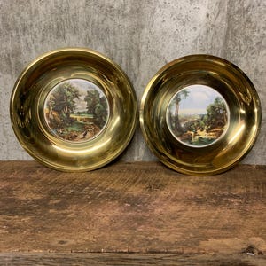 May include: Two brass wall plates with a raised rim and a porcelain centre featuring a landscape scene.