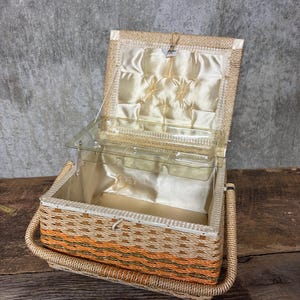 May include: Open vintage wicker sewing basket with a clear plastic tray and a cream-colored satin interior. The basket features orange and brown woven accents and a woven handle.