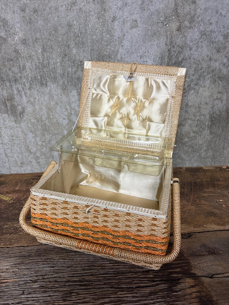 May include: Open vintage wicker sewing basket with a clear plastic tray and a cream-colored satin interior. The basket features orange and brown woven accents and a woven handle.