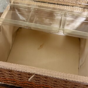 May include: Open vintage wicker sewing basket with a tan interior and clear plastic trays. The basket has a woven design in shades of orange, tan, and brown, with a cream-colored trim and a white closure.