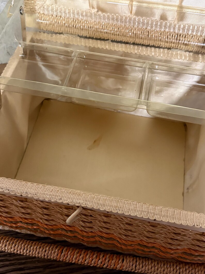 May include: Open vintage wicker sewing basket with a tan interior and clear plastic trays. The basket has a woven design in shades of orange, tan, and brown, with a cream-colored trim and a white closure.