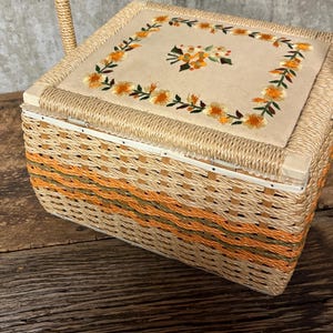 May include: A vintage sewing basket with a woven wicker design in shades of tan, orange, and green. The lid features a beige fabric panel with embroidered orange flowers and green leaves. A woven handle is attached.