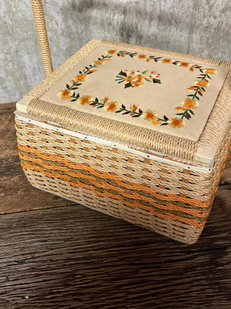 May include: A vintage sewing basket with a woven wicker design in shades of tan, orange, and green. The lid features a beige fabric panel with embroidered orange flowers and green leaves. A woven handle is attached.